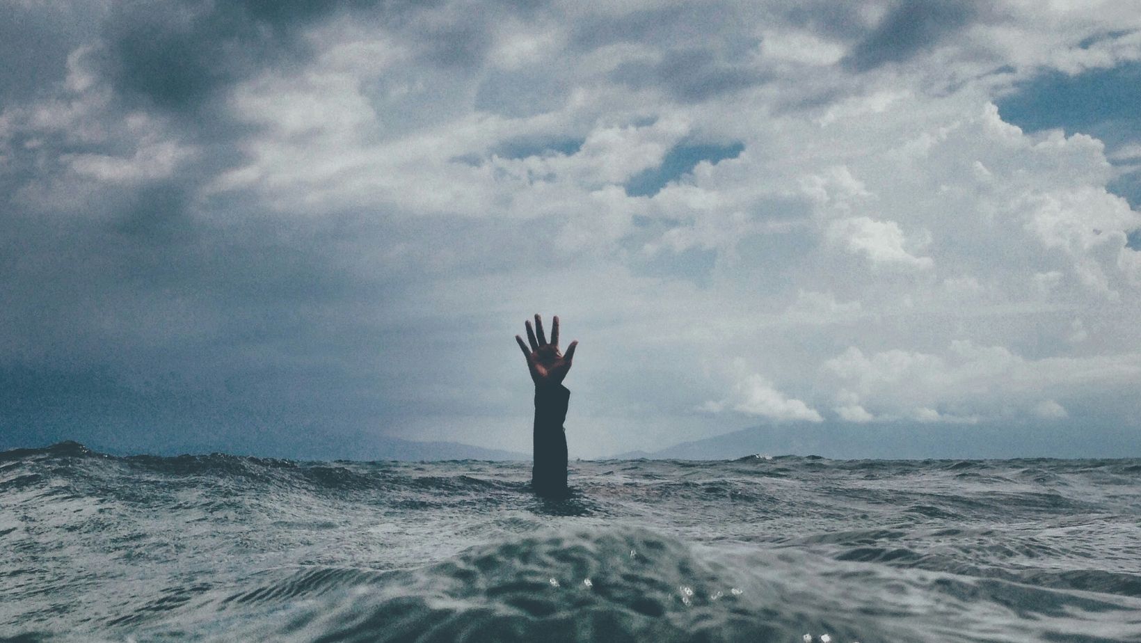A single hand reaching out of turbulent ocean water under a cloudy sky, symbolizing burnout.