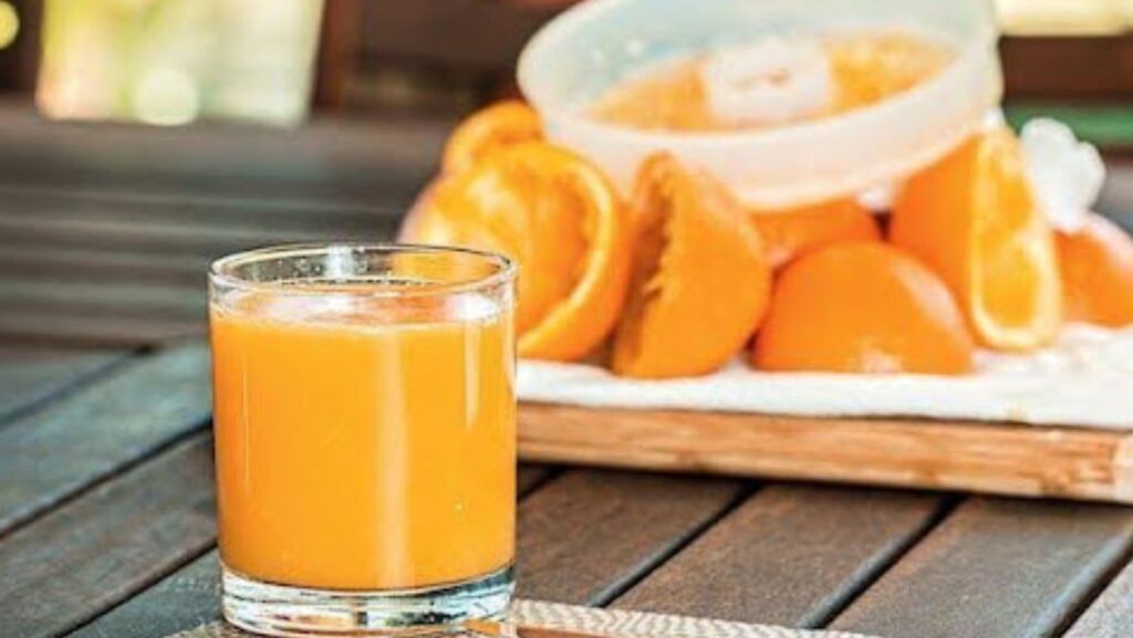 A glass of freshly squeezed orange juice on a wooden table, with squeezed oranges visible in the background.