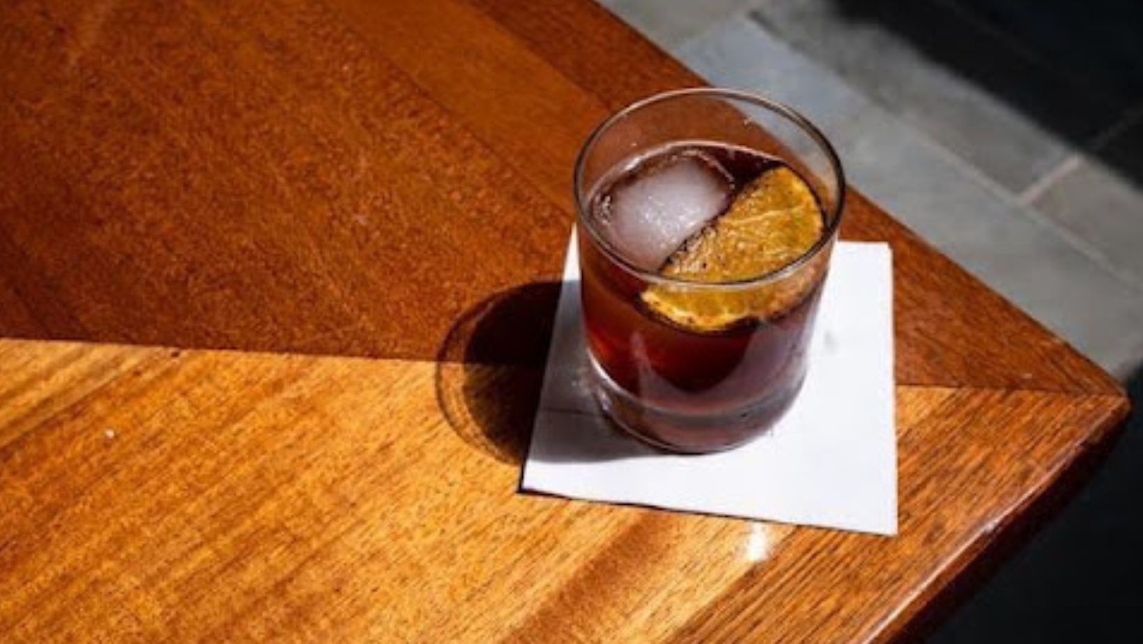 A dark cocktail with ice and an orange slice on a white napkin, on the corner of a wooden bar counter.